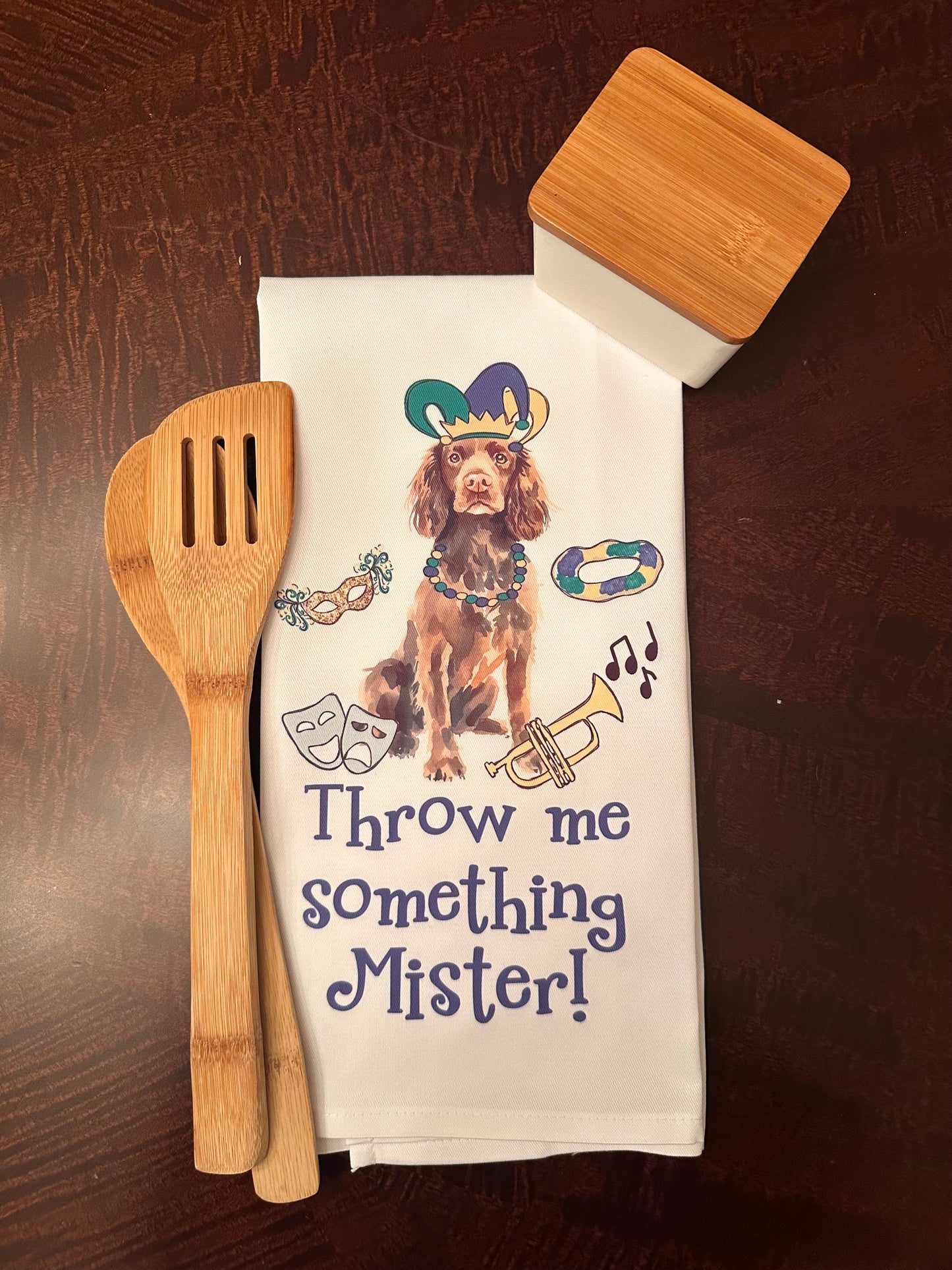 Mardi Gras-themed kitchen towel featuring a festive dog illustration wearing a jester hat and colorful beads, surrounded by carnival elements such as a mask, king cake, trumpet, and musical notes. Text reads 'Throw me something, Mister!' displayed alongside bamboo utensils and a modern storage container on a wooden surface.