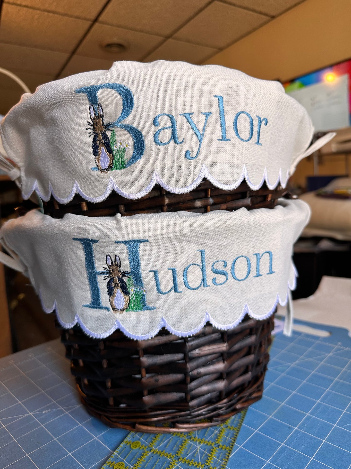 Personalized embroidered Easter basket liners featuring Peter Rabbit design and custom names 'Baylor' and 'Hudson' in soft blue thread, displayed on woven brown baskets with scalloped white trim – custom Easter baskets for kids.