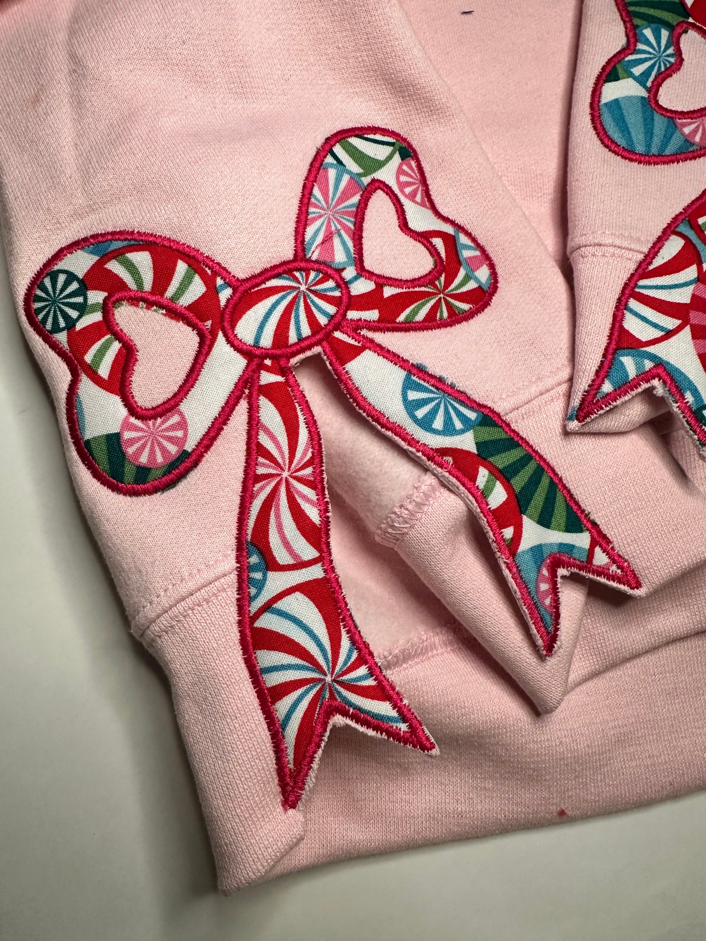 Light pink sweatshirt with festive appliqué bows on the sides, featuring red, green, and blue candy-patterned fabric.