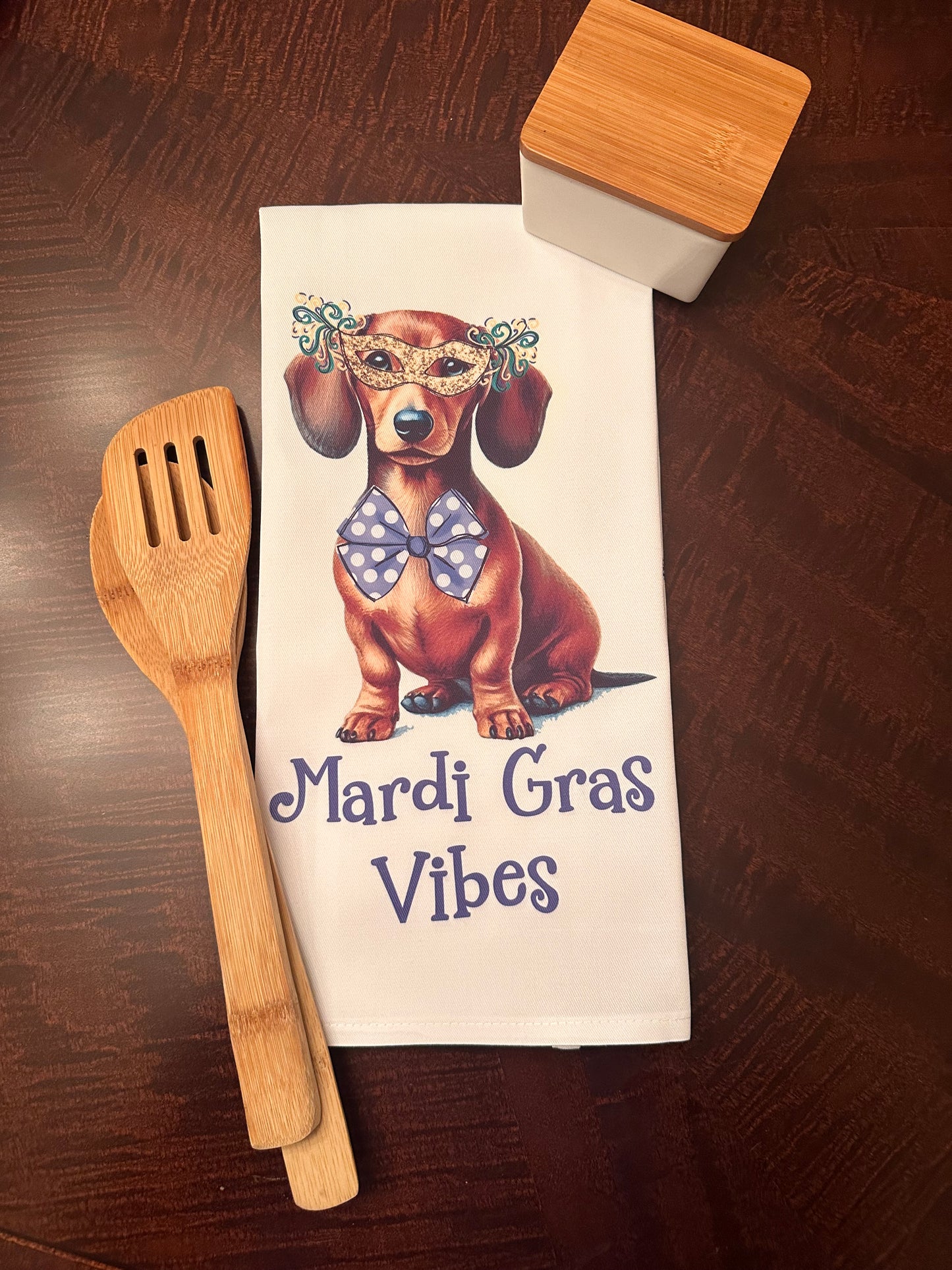 Mardi Gras-themed kitchen towel featuring a dachshund illustration wearing a decorative mask and polka dot bow tie, styled with bamboo utensils and a modern storage container on a wooden table.