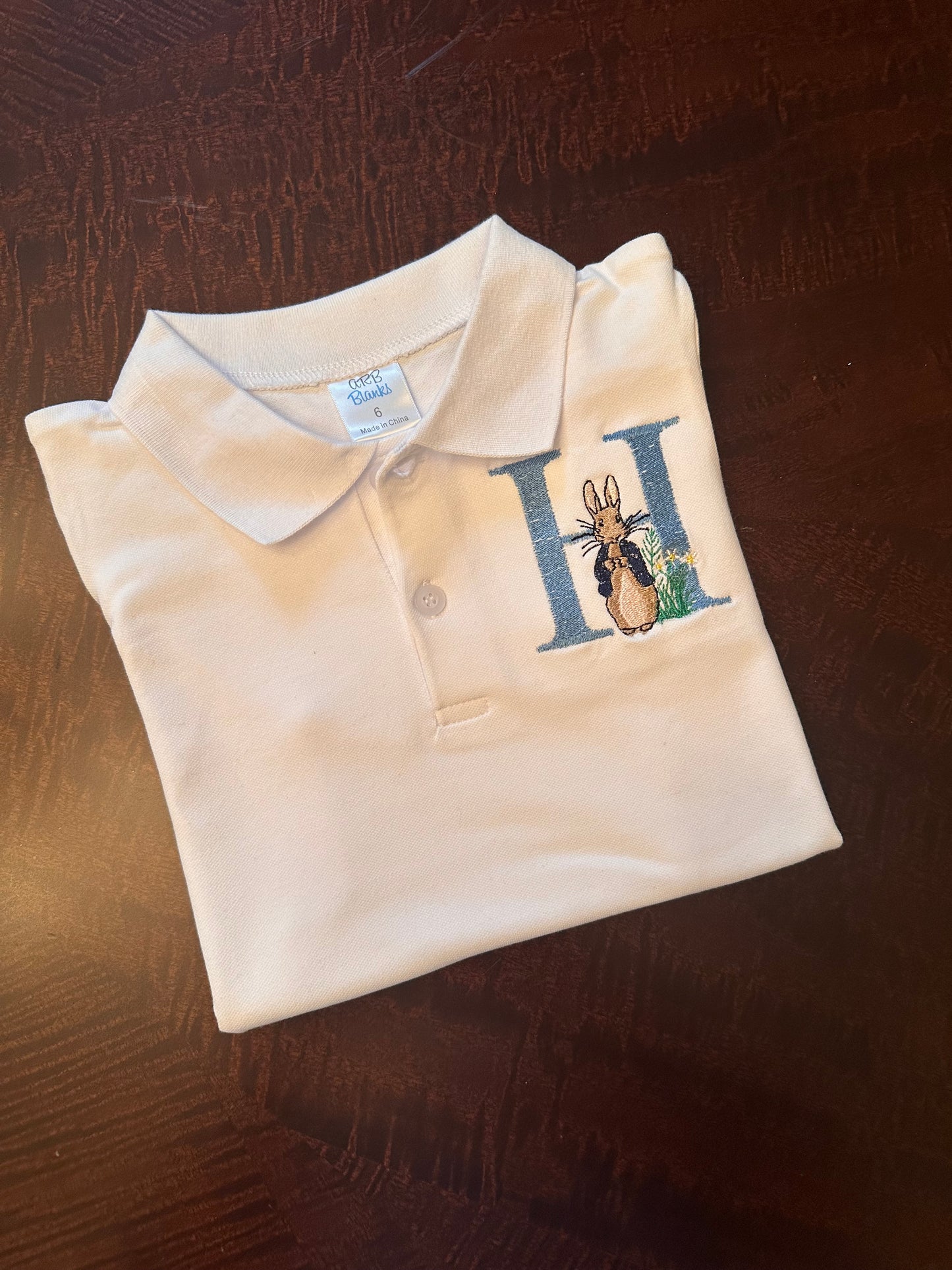 Personalized boys Easter polo shirt with blue embroidered initial 'H' and Peter Rabbit design on white collared shirt β custom monogram Easter outfit for toddlers and kids.