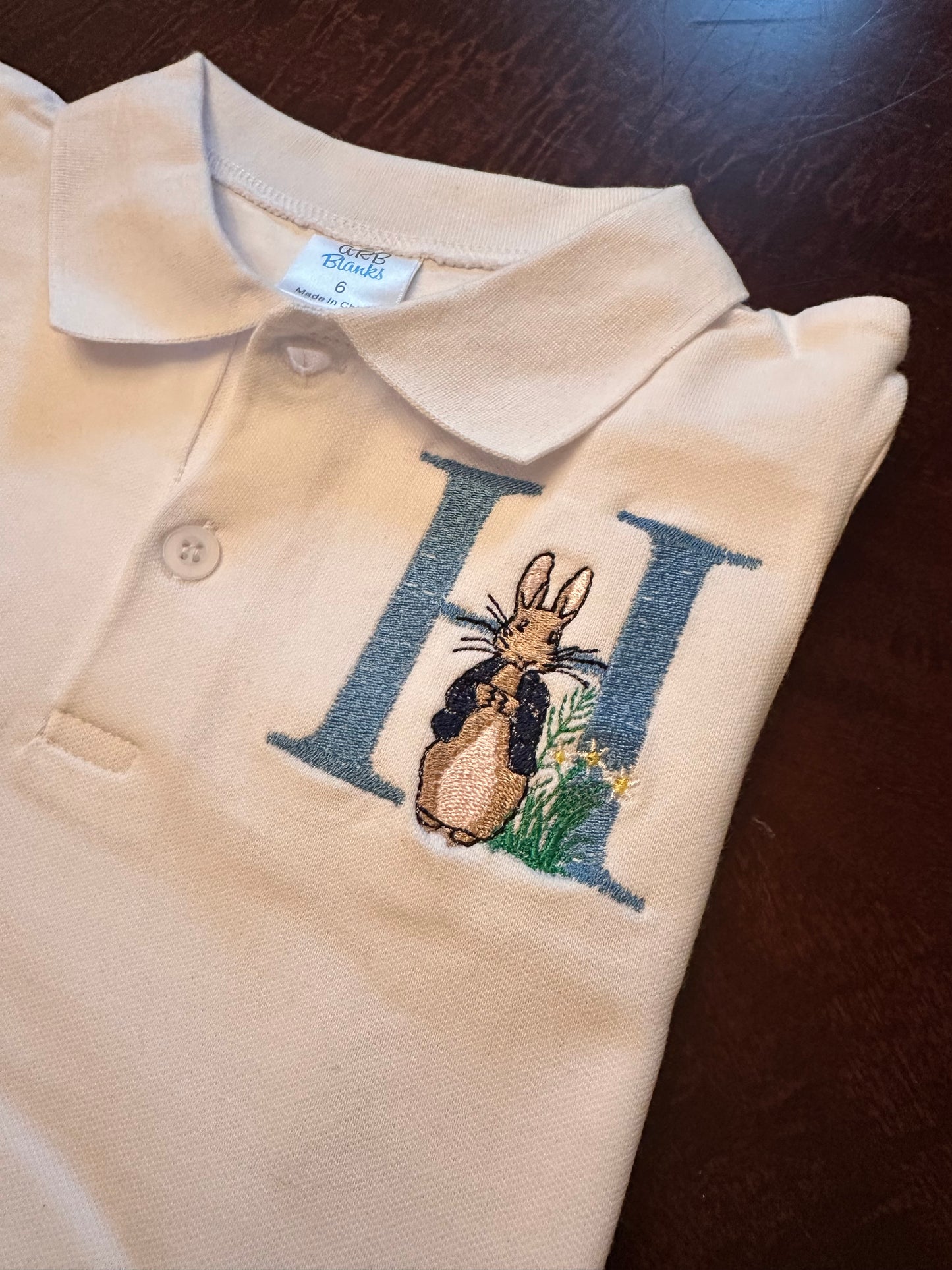 Personalized boys Easter polo shirt with blue embroidered initial 'H' and Peter Rabbit design on white collared shirt β custom monogram Easter outfit for toddlers and kids.