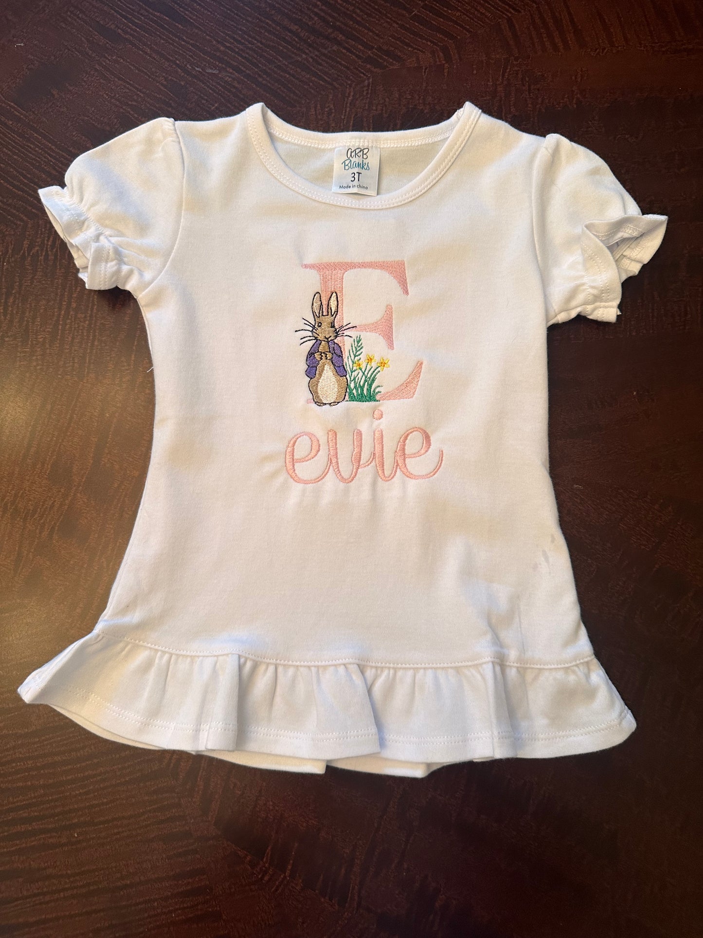Personalized girls Easter shirt with pink embroidered initial 'E', Peter Rabbit design, and custom name 'Evie' on white ruffle hem tee β custom Easter outfit for toddlers and kids.