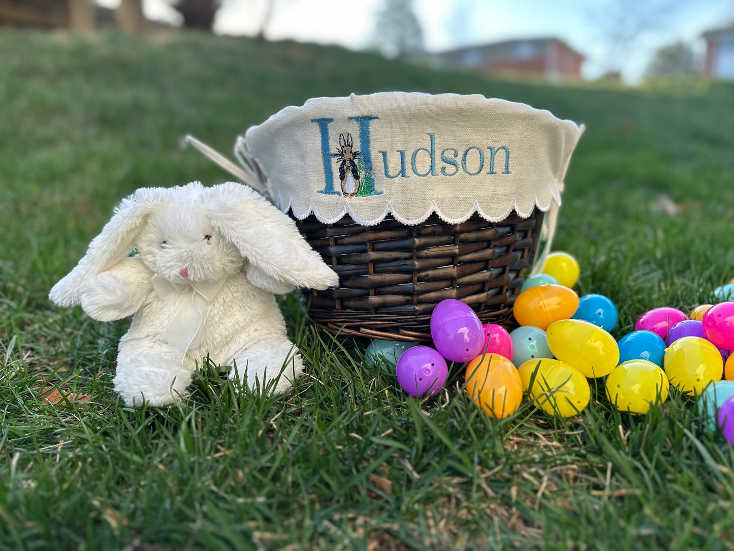 Personalized Easter basket with Peter Rabbit embroidered liner featuring the name 'Hudson,' displayed on grass with colorful plastic Easter eggs and a plush white bunny – custom Easter gift for kids.