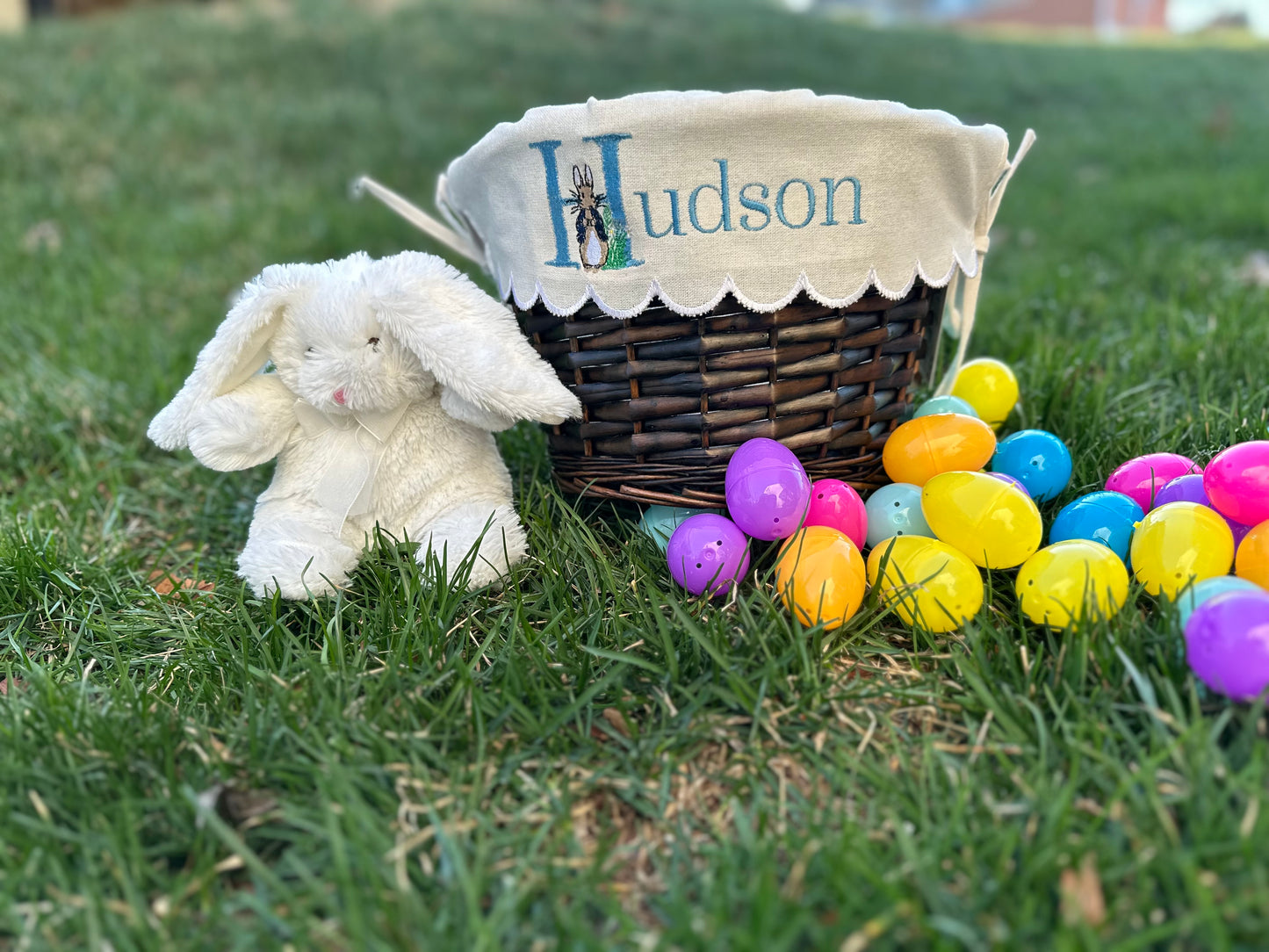 Personalized Easter basket with Peter Rabbit embroidered liner featuring the name 'Hudson,' displayed on grass with colorful plastic Easter eggs and a plush white bunny – custom Easter gift for kids.