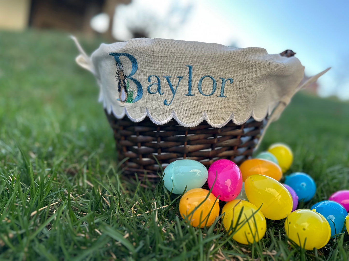 Personalized Easter basket with custom embroidered liner featuring the name 'Baylor' and Peter Rabbit design on scalloped fabric, displayed on grass with colorful plastic Easter eggs – custom Easter basket for kids.