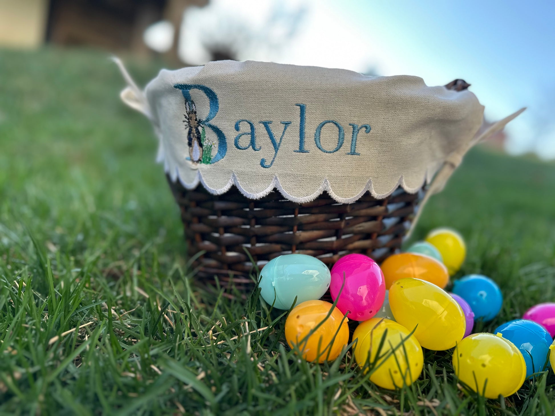 Personalized Easter basket with custom embroidered liner featuring the name 'Baylor' and Peter Rabbit design on scalloped fabric, displayed on grass with colorful plastic Easter eggs – custom Easter basket for kids.