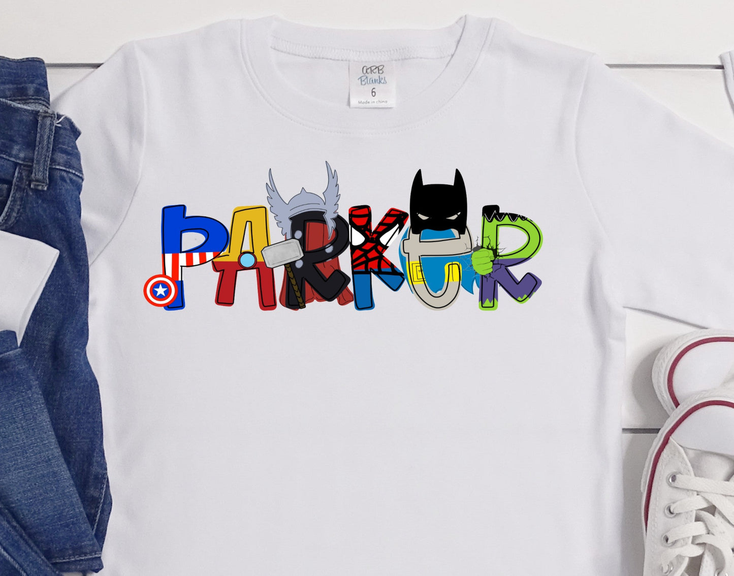 White t-shirt with colorful text design of letters with various superhero designs, blue jeans, and white sneakers on a light wooden surface