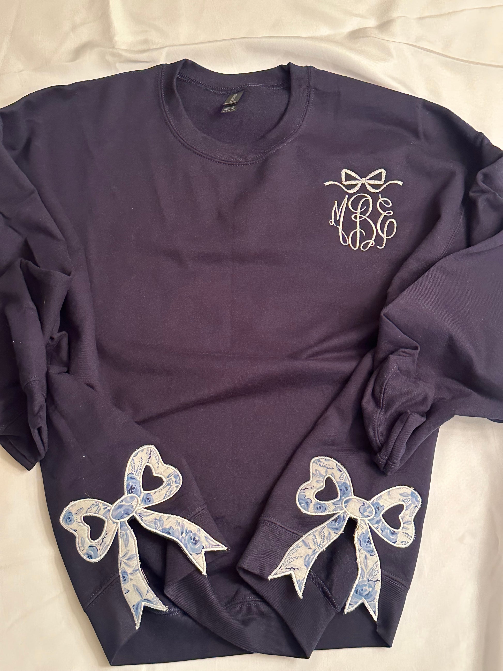 Navy sweatshirt with white bow detail  and monogram on the chest, featuring two blue and white floral heart-cutout side bow appliqués on each side near the hem.