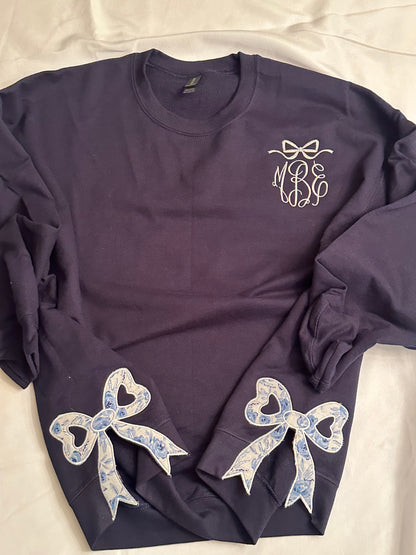 Navy sweatshirt with white bow detail  and monogram on the chest, featuring two blue and white floral heart-cutout side bow appliqués on each side near the hem.