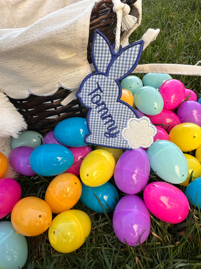 Personalized embroidered Easter basket tag in blue gingham bunny shape with the name 'Tommy,' hanging on a woven Easter basket surrounded by colorful plastic Easter eggs on grass.