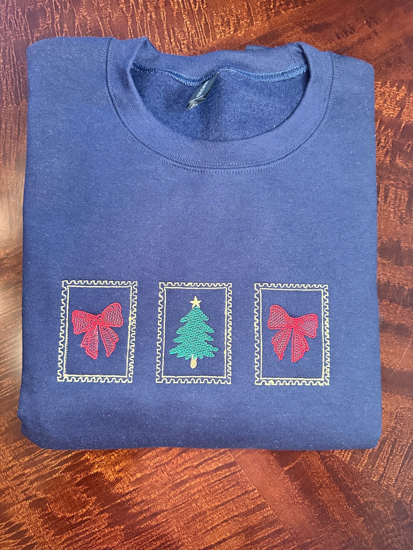 Folded navy sweatshirt with embroidered Christmas design — red bows and green Christmas tree framed in gold.