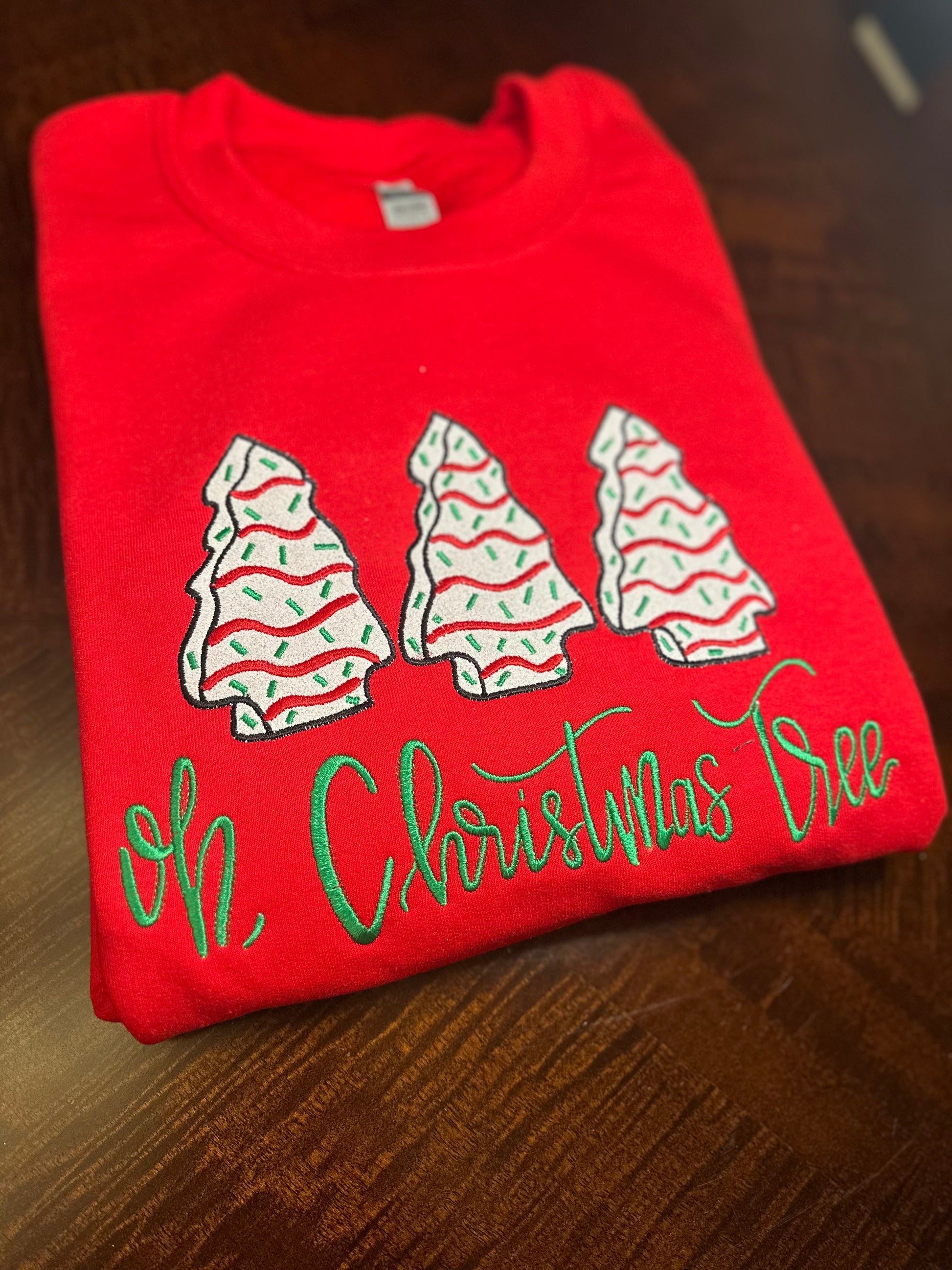 Folded red sweatshirt with embroidered Christmas trees and green “Oh, Christmas Tree” text.
