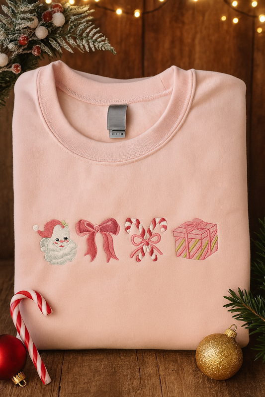 Folded pink sweatshirt with embroidered Christmas icons — Santa, bow, candy canes, and gift box — surrounded by festive holiday decor and lights.