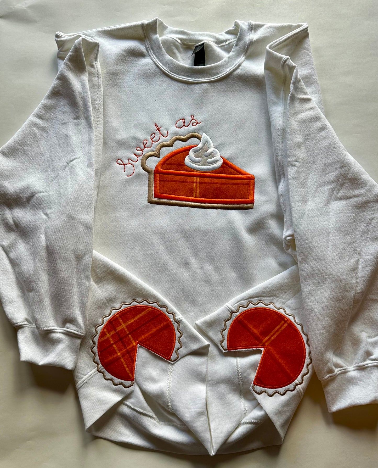 White embroidered crewneck sweatshirt laid flat showing “Sweet as Pie” design with matching pumpkin pie embroidery on sleeves — festive Thanksgiving apparel.