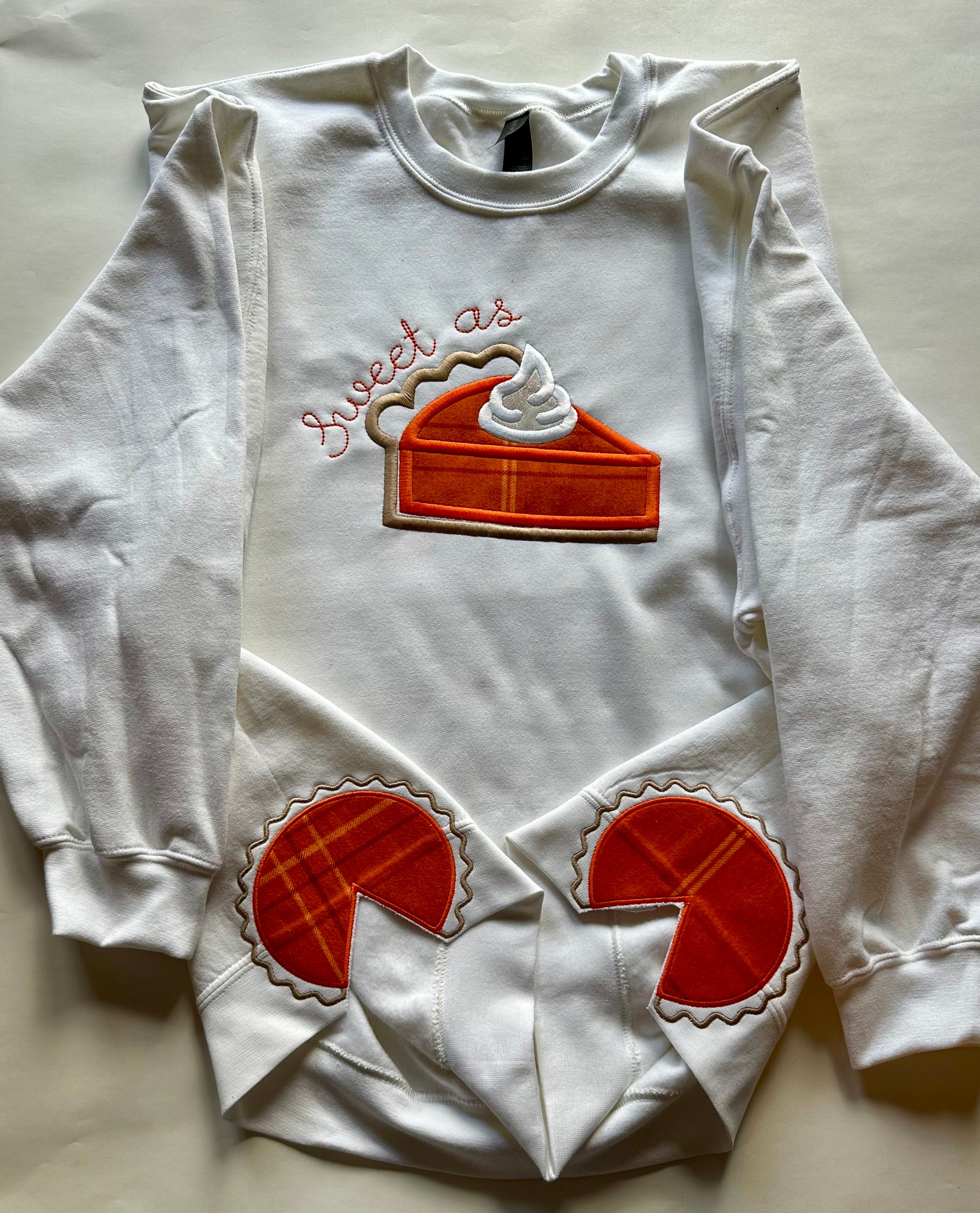 White embroidered crewneck sweatshirt laid flat showing “Sweet as Pie” design with matching pumpkin pie embroidery on sleeves — festive Thanksgiving apparel.
