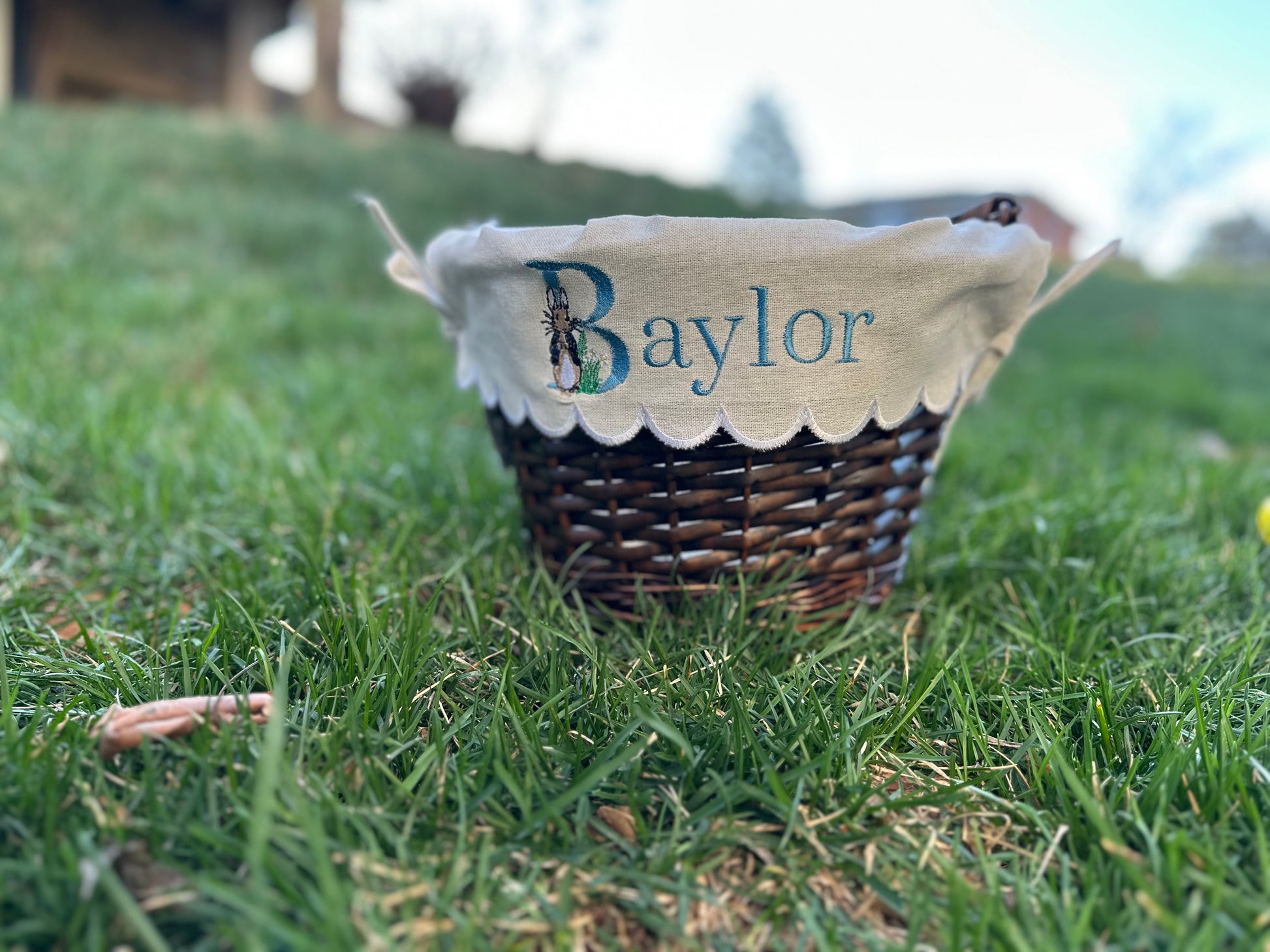 Custom embroidered Easter basket liner featuring Peter Rabbit design and personalized name 'Baylor' in blue thread on scalloped white fabric, displayed on a wicker basket set in grass – personalized Easter basket for kids.