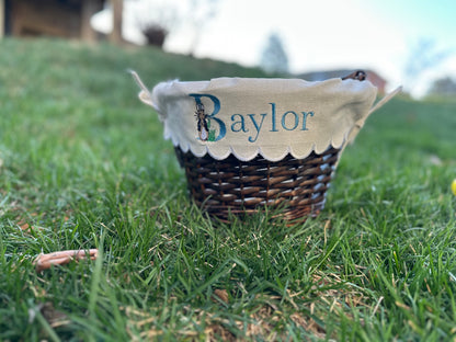 Custom embroidered Easter basket liner featuring Peter Rabbit design and personalized name 'Baylor' in blue thread on scalloped white fabric, displayed on a wicker basket set in grass – personalized Easter basket for kids.
