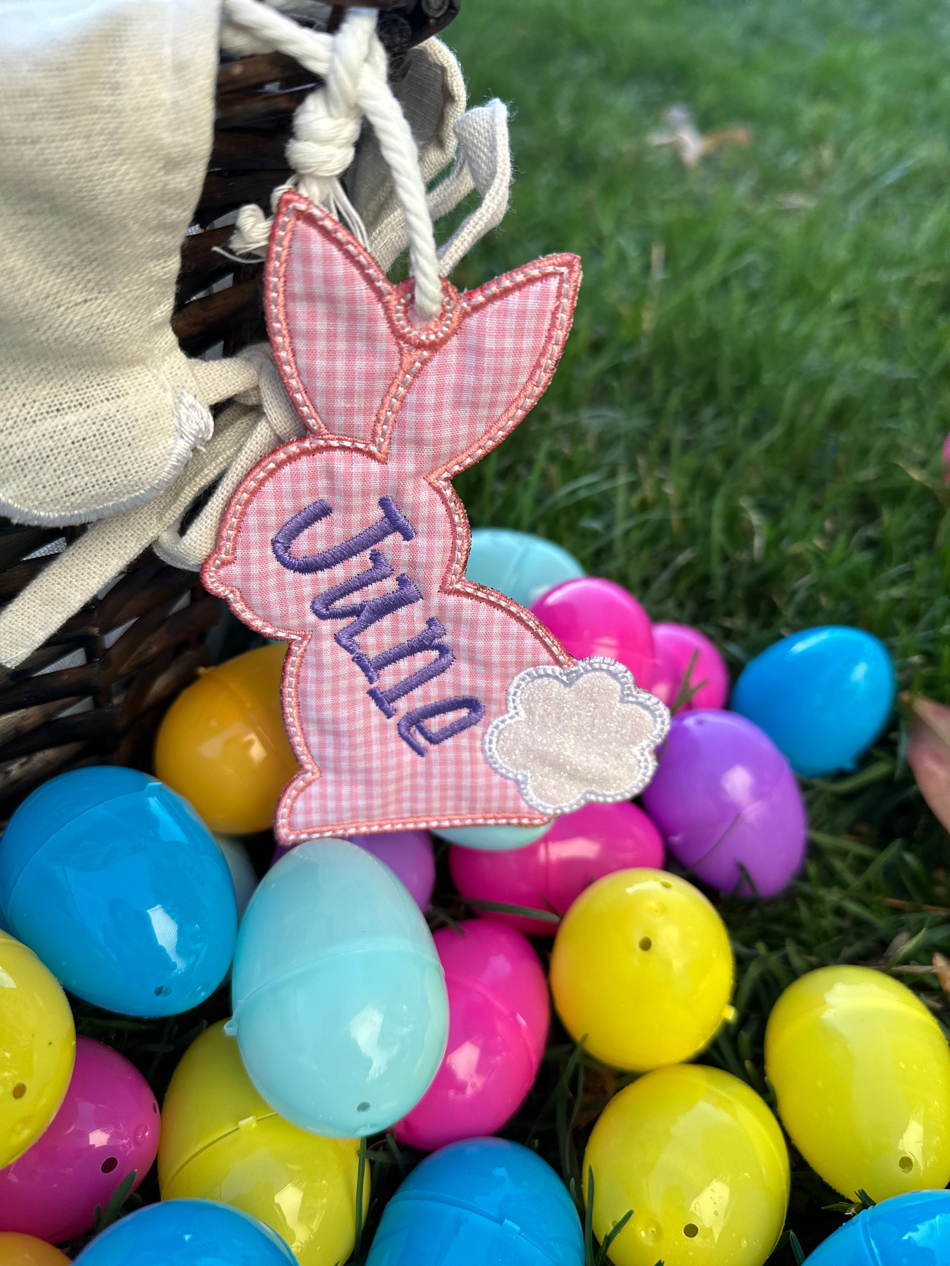 Custom embroidered pink gingham bunny-shaped Easter basket tag with name 'June,' featuring a white glitter tail, attached to a wicker basket surrounded by colorful plastic Easter eggs – personalized Easter gift for toddlers and kids.