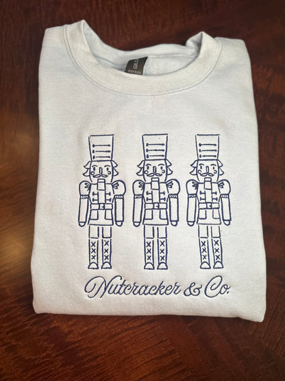 Folded light blue and white embroidered sweatshirts featuring three nutcrackers and “Nutcracker & Co.” script.