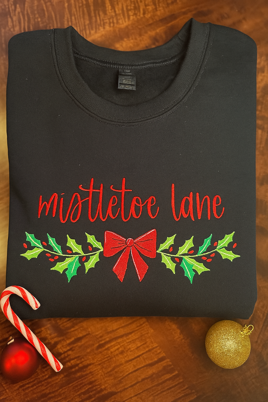 Folded black sweatshirt with embroidered red “mistletoe lane” text and green holly leaves with a red bow, styled with festive Christmas decorations including a candy cane and gold ornament.