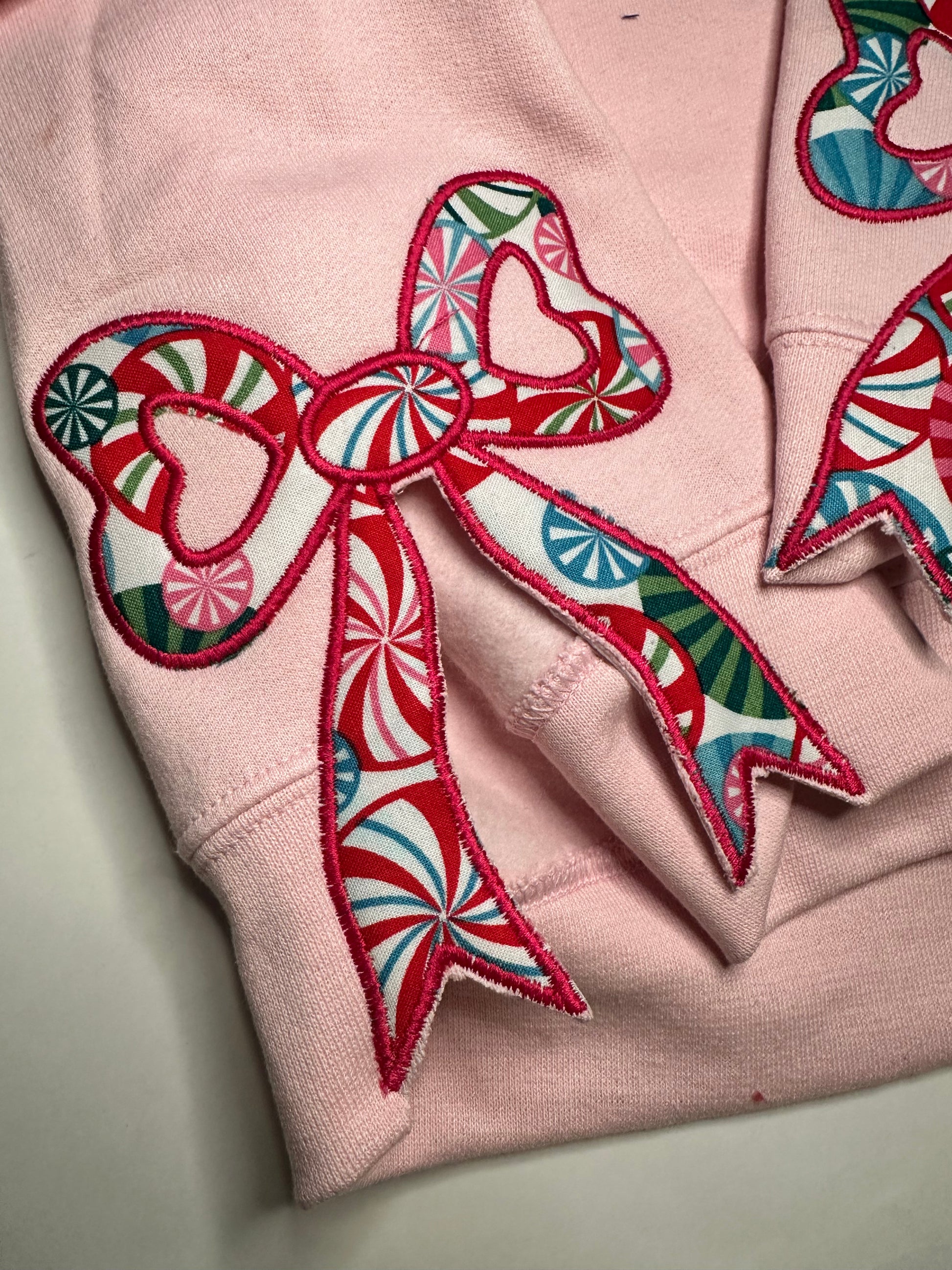 Light pink sweatshirt with festive appliqué bows on the sides, featuring red, green, and blue candy-patterned fabric.