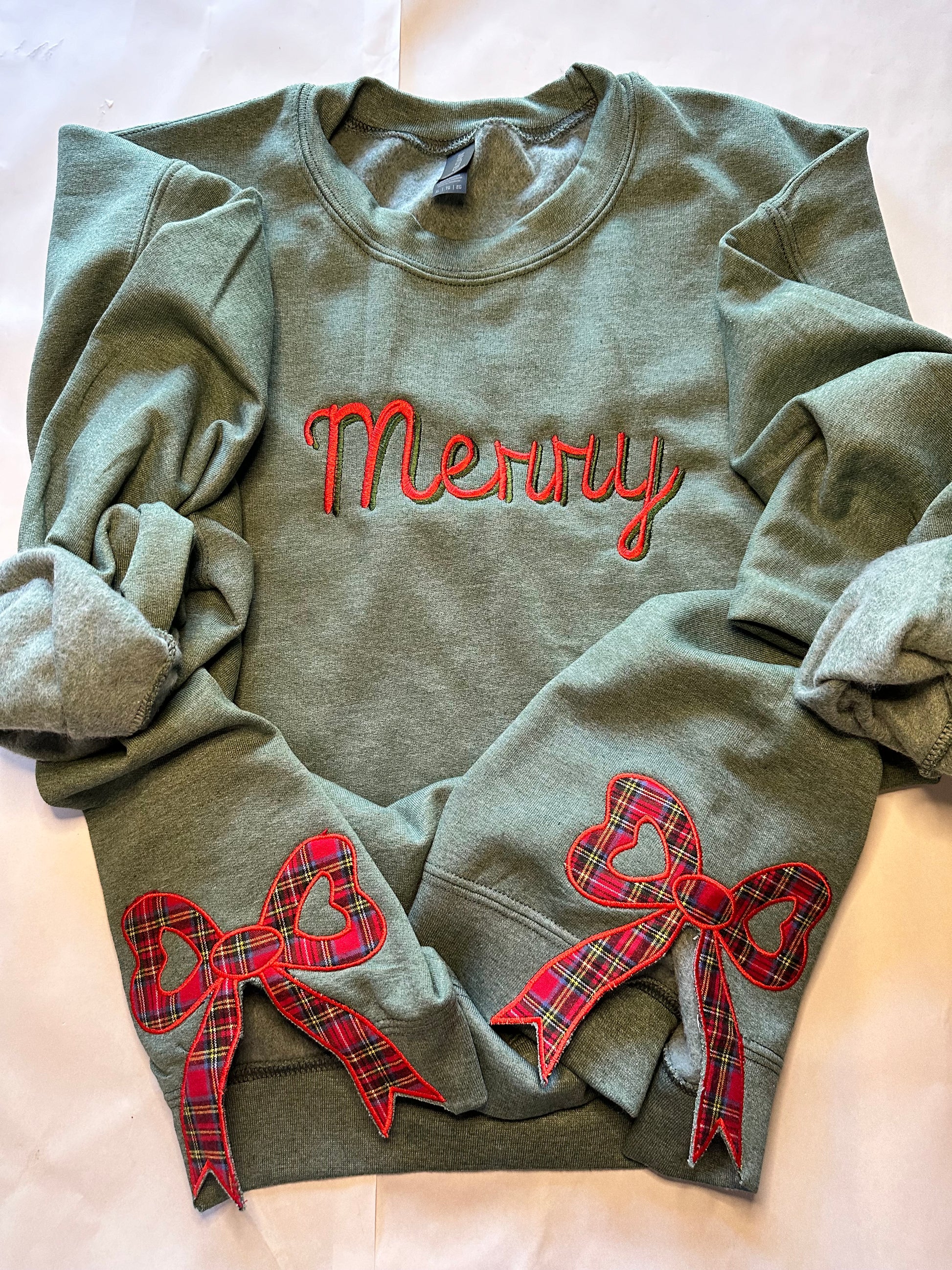 Green sweatshirt with “Merry” embroidered in red and green across the chest and two red plaid side bow appliqués with heart-shaped centers, displayed on a light background.
