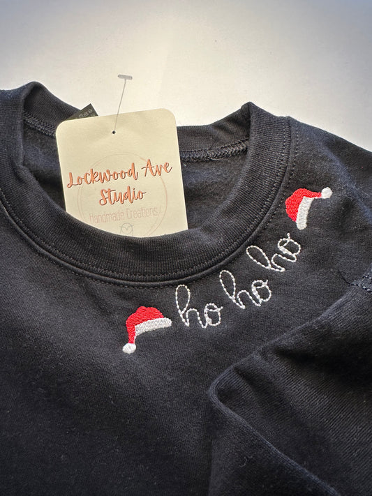 Children’s black sweatshirt featuring “ho ho ho” embroidery with Santa hats on the neckline 