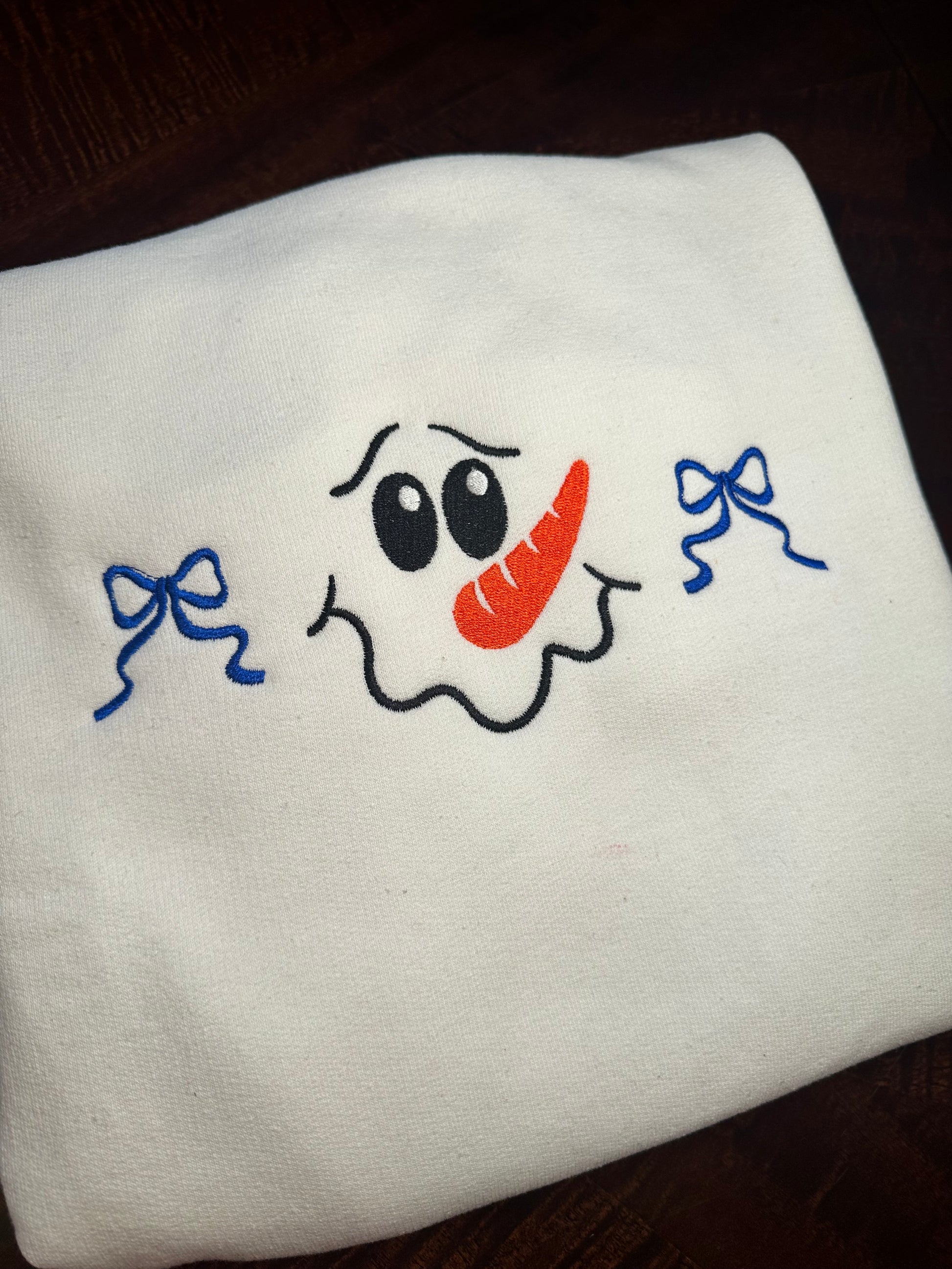 Folded white sweatshirt with embroidered snowman face featuring a carrot nose, black eyes, and two blue bows.