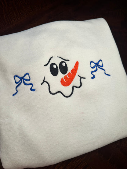 Folded white sweatshirt with embroidered snowman face featuring a carrot nose, black eyes, and two blue bows.
