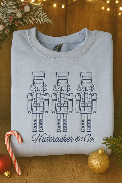 Folded light blue and white embroidered sweatshirts featuring three nutcrackers and “Nutcracker & Co.” script, styled with festive Christmas decor including candy canes, ornaments, and twinkling lights.