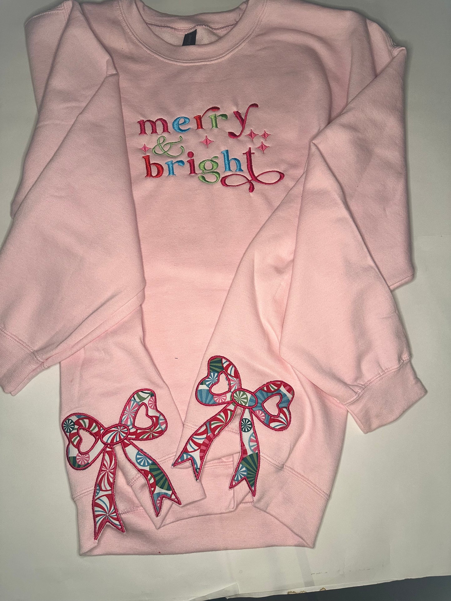Light pink sweatshirt with colorful “merry & bright” embroidery on the chest and two festive appliqué bows on the sides, featuring red, green, and blue candy-patterned fabric.