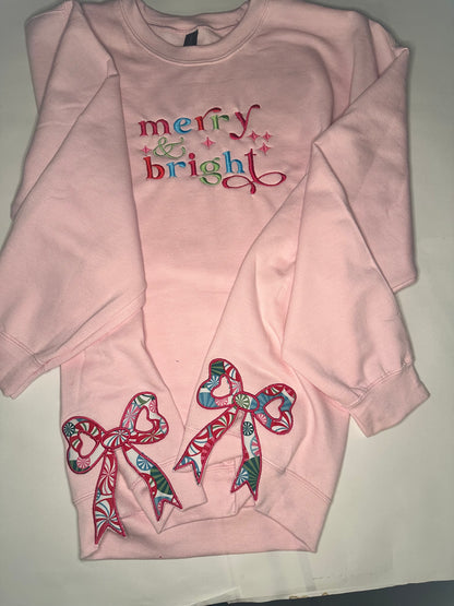 Light pink sweatshirt with colorful “merry & bright” embroidery on the chest and two festive appliqué bows on the sides, featuring red, green, and blue candy-patterned fabric.