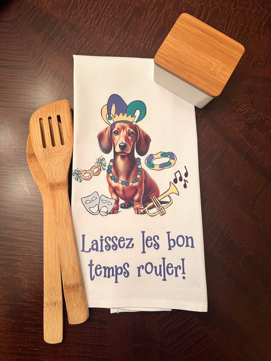 Mardi Gras kitchen towel featuring a dachshund illustration wearing a jester hat, colorful beads, and surrounded by festive elements like a mask, king cake, and musical notes, paired with bamboo utensils and a modern storage container on a wooden surface. Text reads 'Laissez les bon temps rouler!'