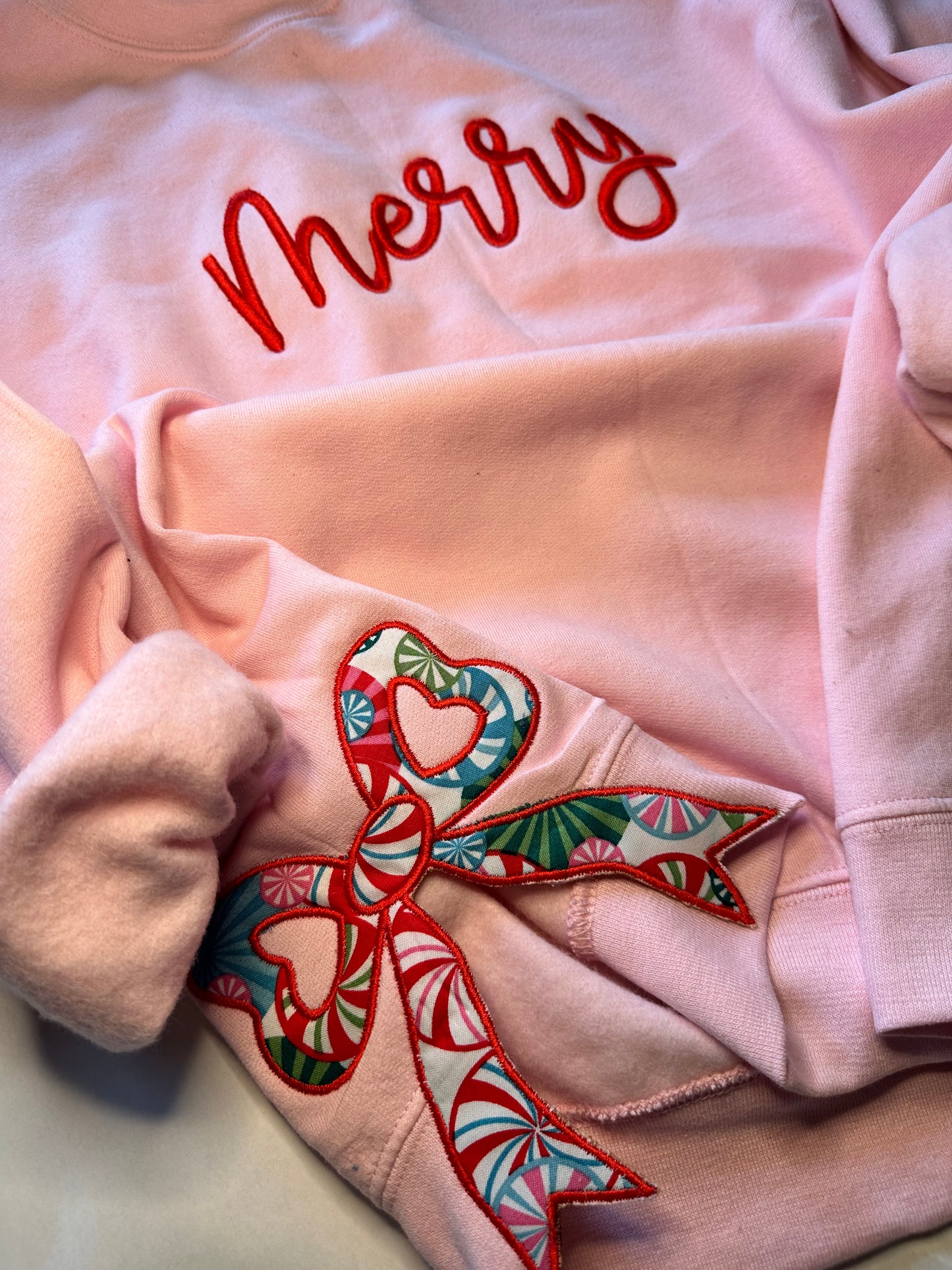 Light pink sweatshirt with red “Merry” embroidery across the chest and two festive patterned side bow appliqués with heart-shaped centers in red, green, and blue holiday prints.