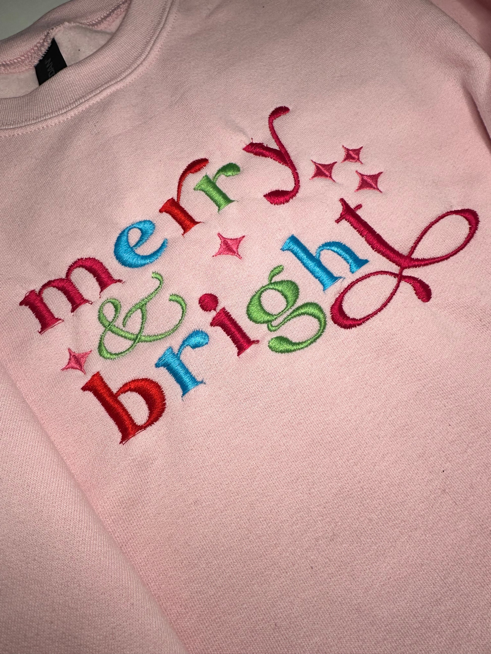Embroidered Merry and Bright wording in red, pink, blue and green colors on a light pink crewneck sweatshirt