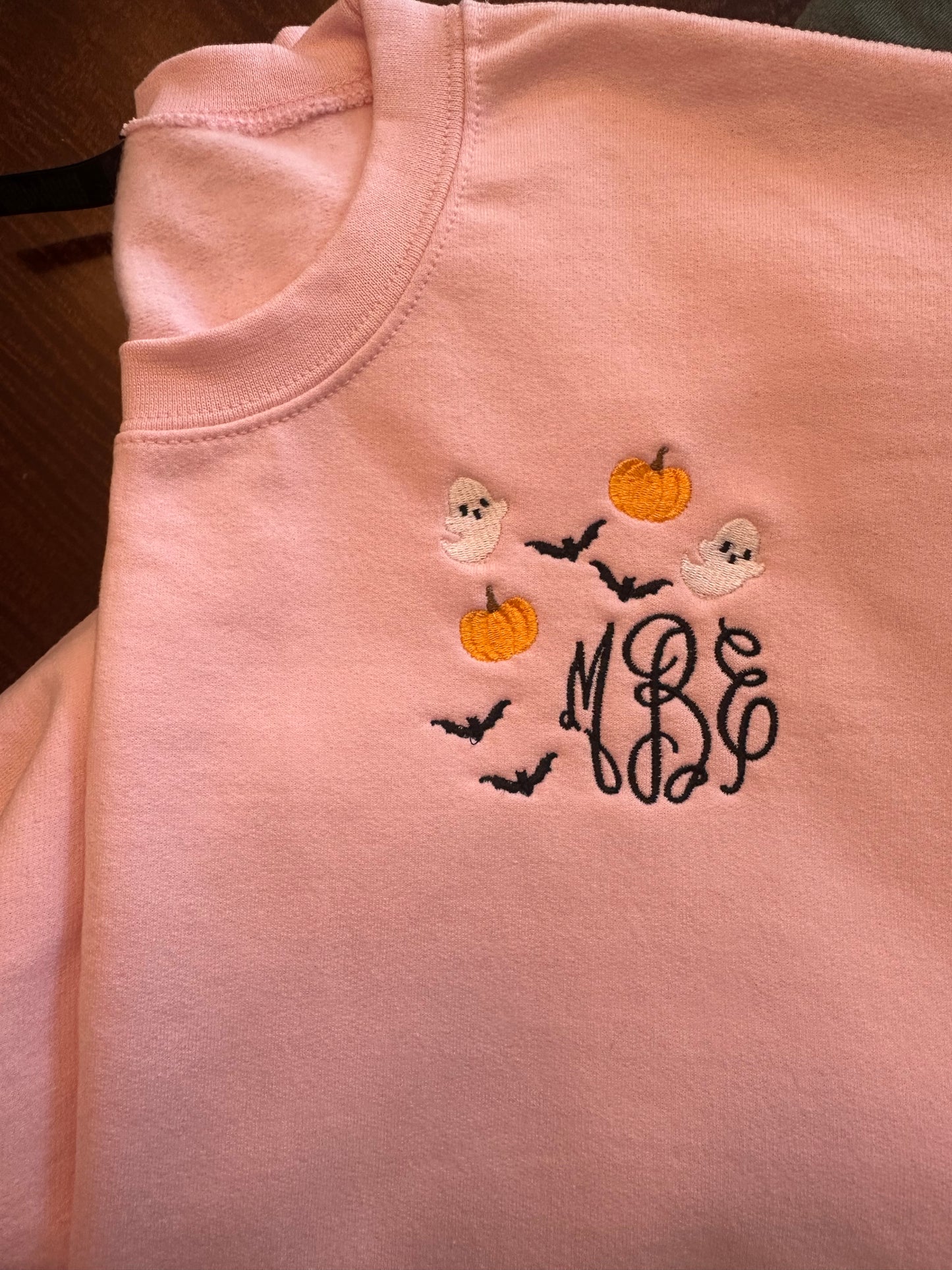 Light pink embroidered crewneck sweatshirt with black monogram surrounded by small pumpkins, ghosts, and bats — custom Halloween sweatshirt