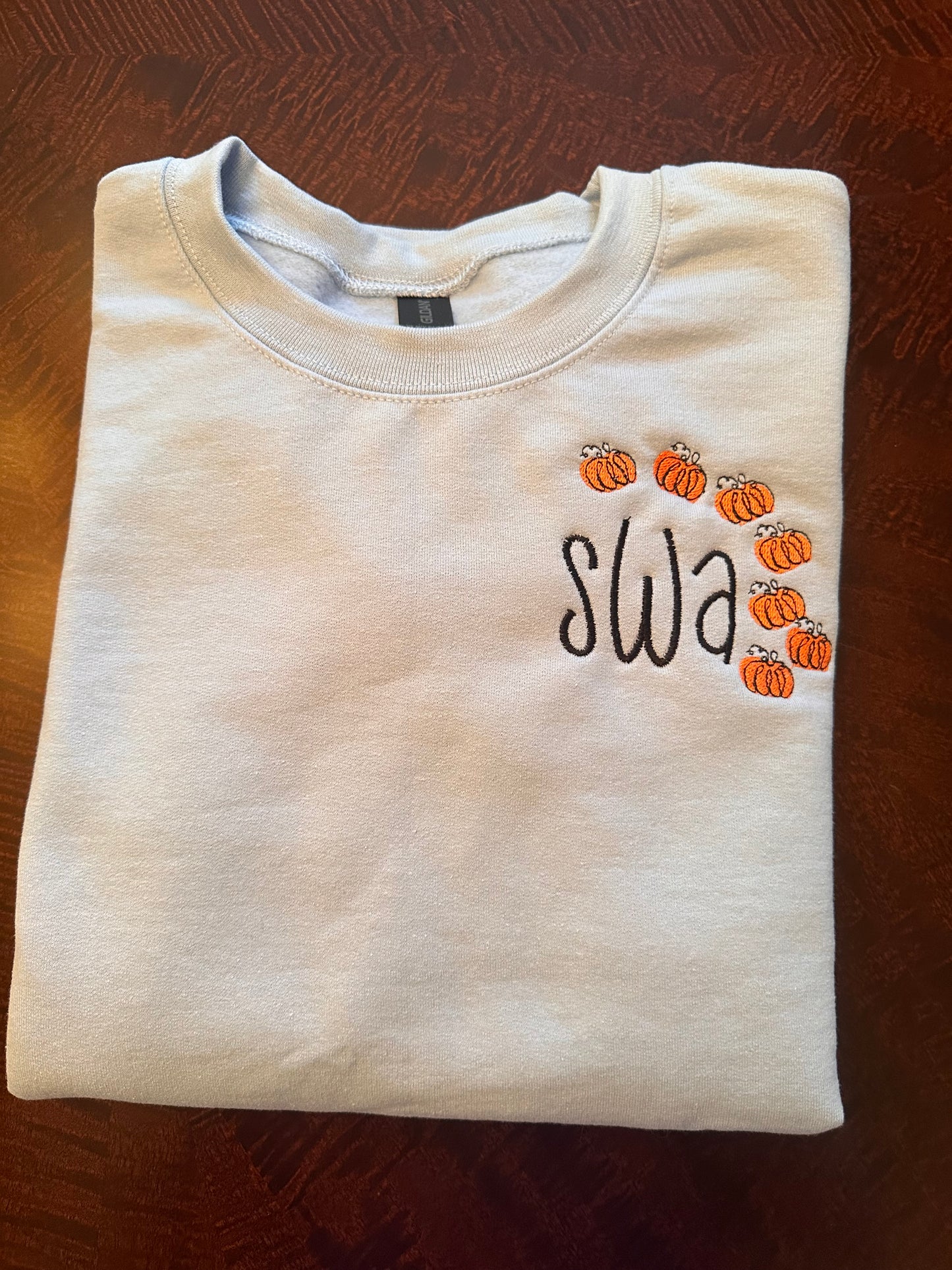 Light blue embroidered crewneck sweatshirt with black monogram letters and small orange pumpkin embroidery frame — personalized fall sweatshirt for women