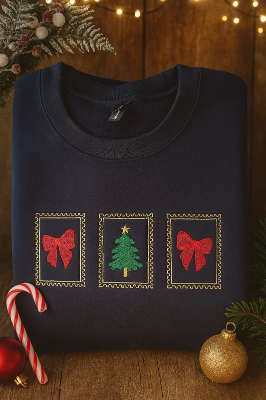 Folded navy sweatshirt with embroidered Christmas design — red bows and green Christmas tree framed in gold — surrounded by festive holiday lights, candy cane, and ornaments.