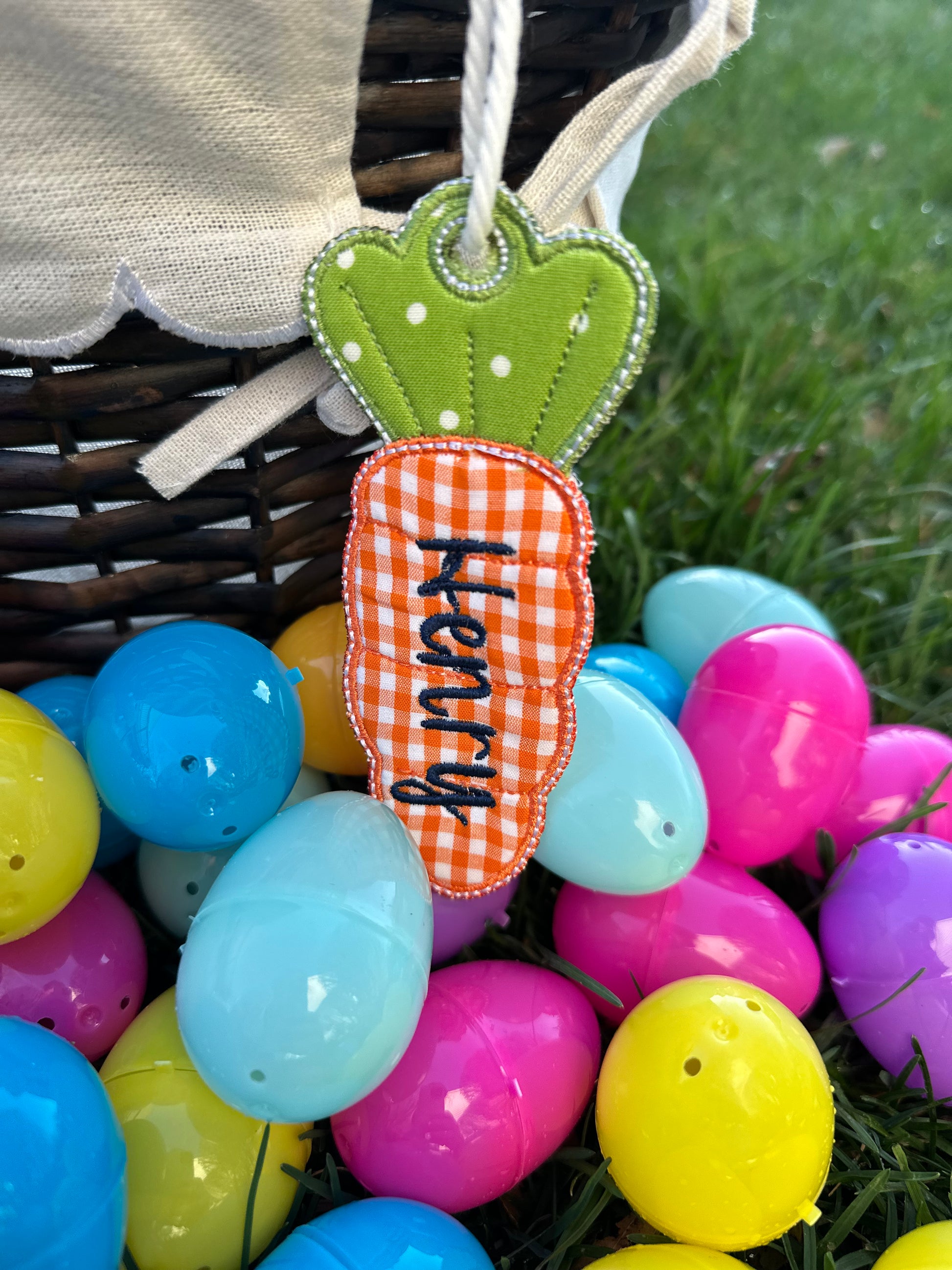 Custom embroidered Easter basket tag in orange gingham carrot shape with the name 'Henry,' attached to a woven basket surrounded by colorful plastic Easter eggs on grass.
