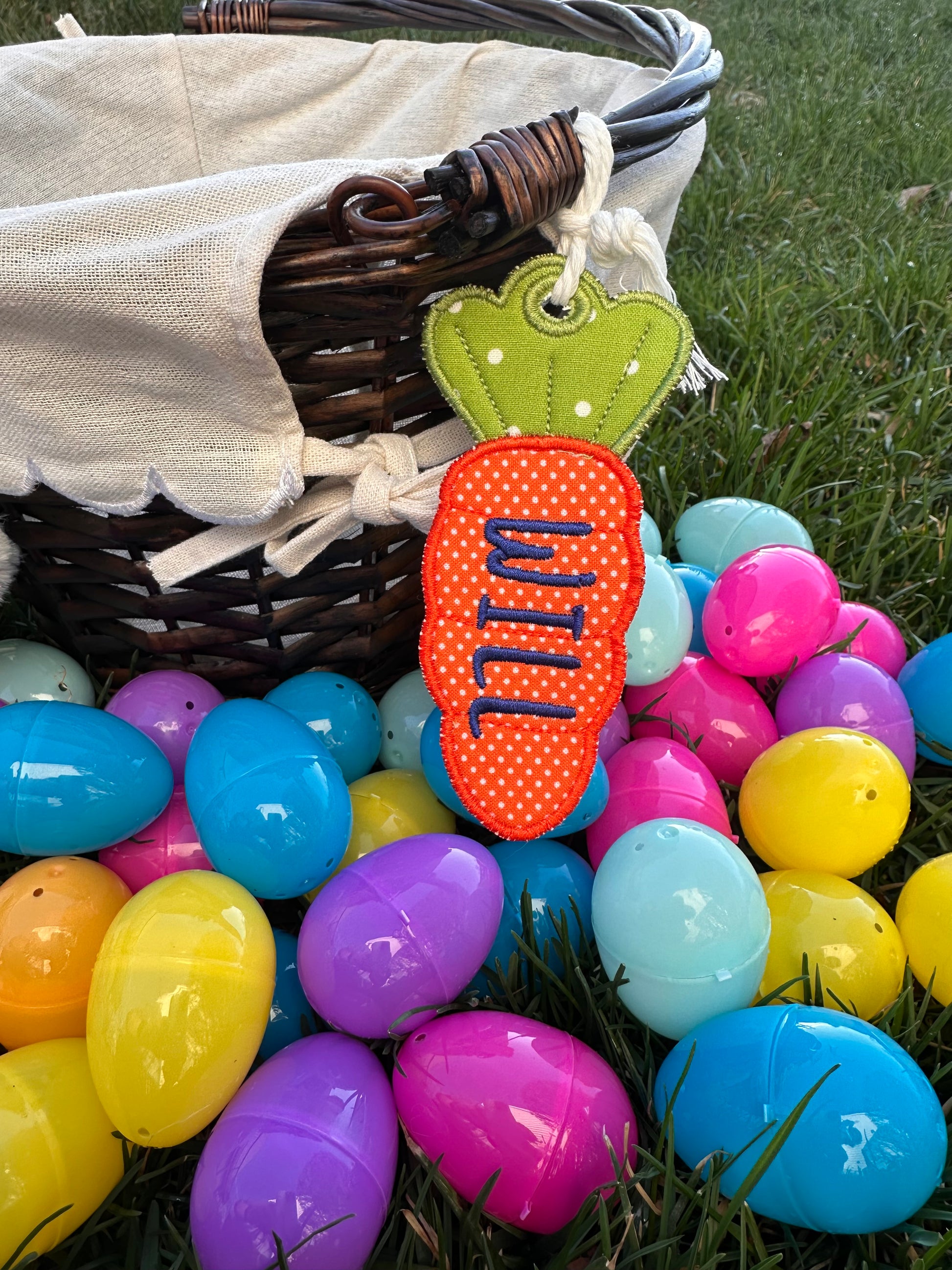 Custom embroidered Easter basket tag in orange swiss dot carrot shape with the name 'Will,' attached to a woven basket surrounded by colorful plastic Easter eggs on grass.