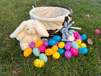Personalized Easter basket with blue gingham bunny name tag labeled 'Tommy,' surrounded by colorful plastic eggs and a plush white bunny, displayed on green grass.