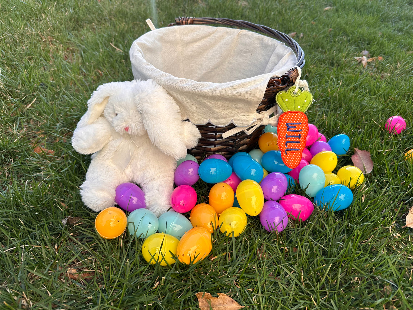 Personalized Easter basket with embroidered carrot name tag labeled 'Will,' surrounded by colorful plastic eggs and a plush white bunny, displayed on green grass.