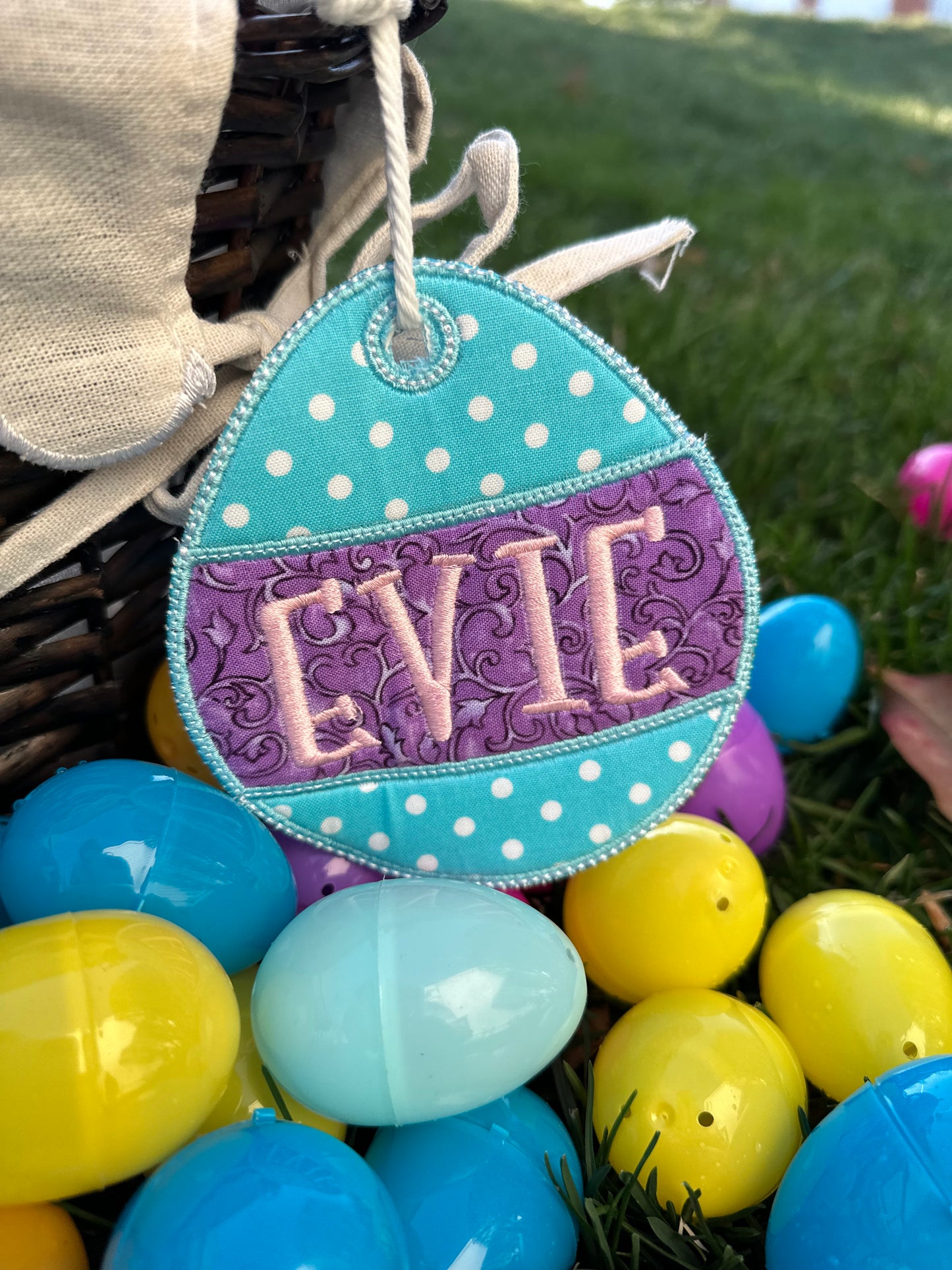 Personalized embroidered Easter basket tag in turquoise and purple polka dot egg shape with custom name 'Evie,' attached to a wicker basket surrounded by colorful plastic Easter eggs – custom Easter gift for kids.