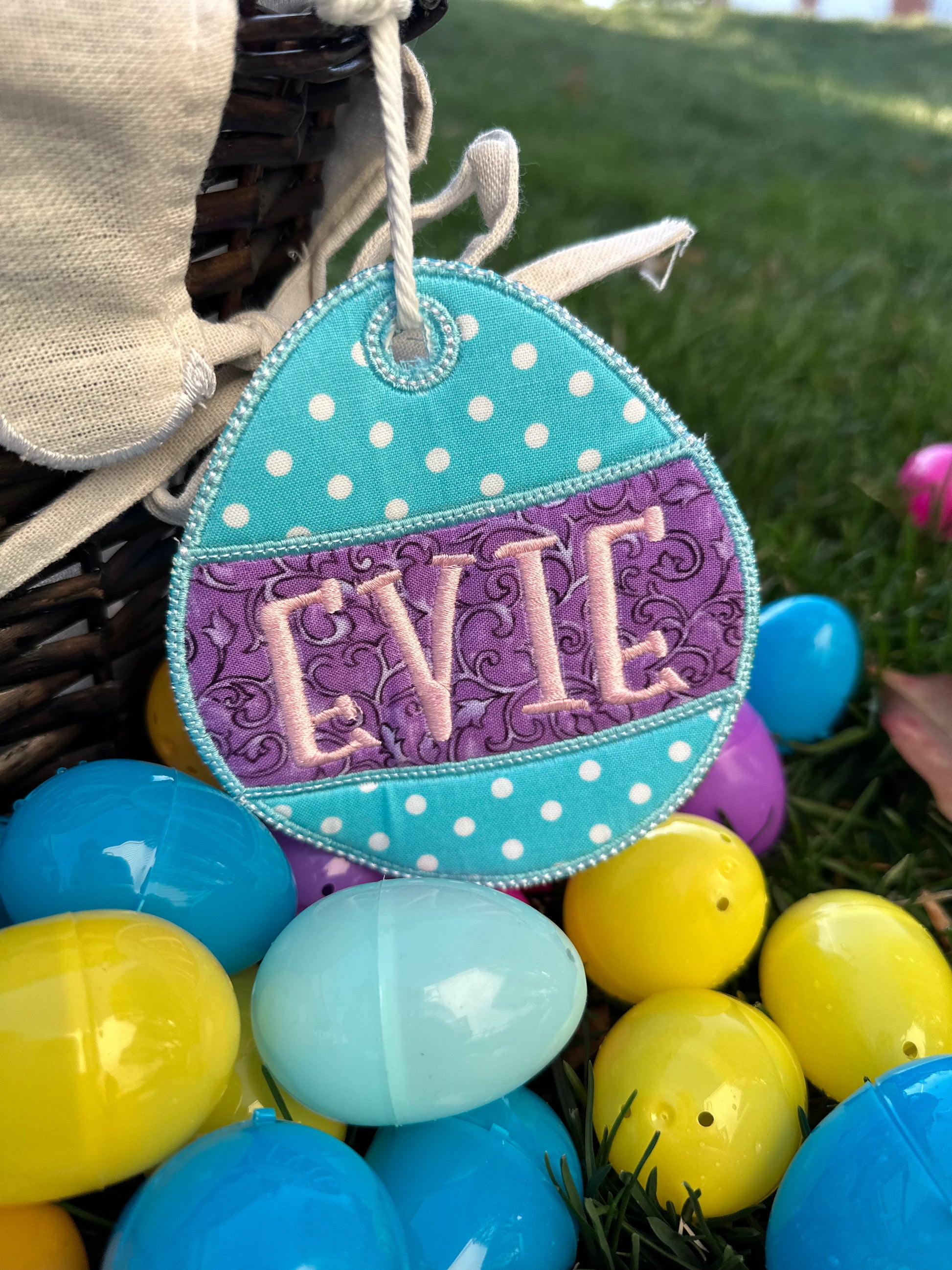 Personalized embroidered Easter basket tag in turquoise and purple polka dot egg shape with custom name 'Evie,' attached to a wicker basket surrounded by colorful plastic Easter eggs – custom Easter gift for kids.