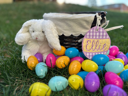 Personalized Easter basket with embroidered egg shaped tag labeled 'Ellie'. with plastic eggs and a plush white bunny, displayed on green grass.