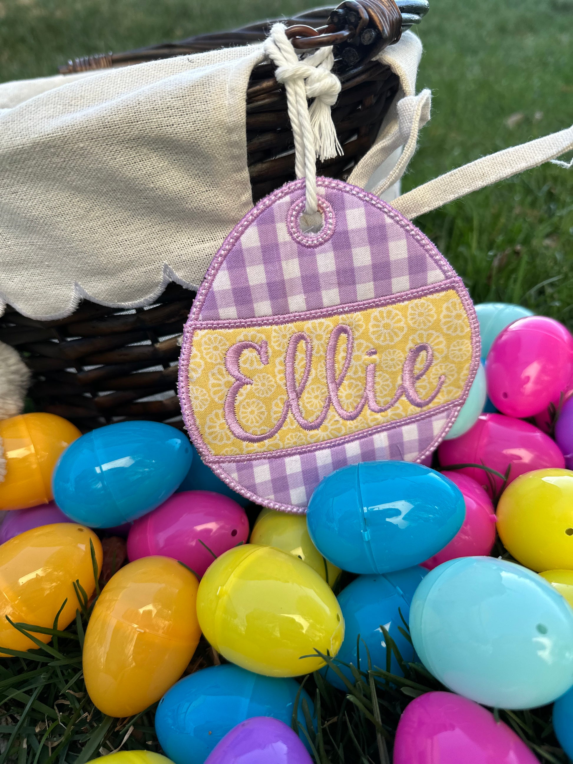 Custom embroidered Easter basket tag in a purple and yellow gingham egg shape with the name 'Ellie,' displayed on a wicker basket surrounded by colorful plastic Easter eggs on grass.