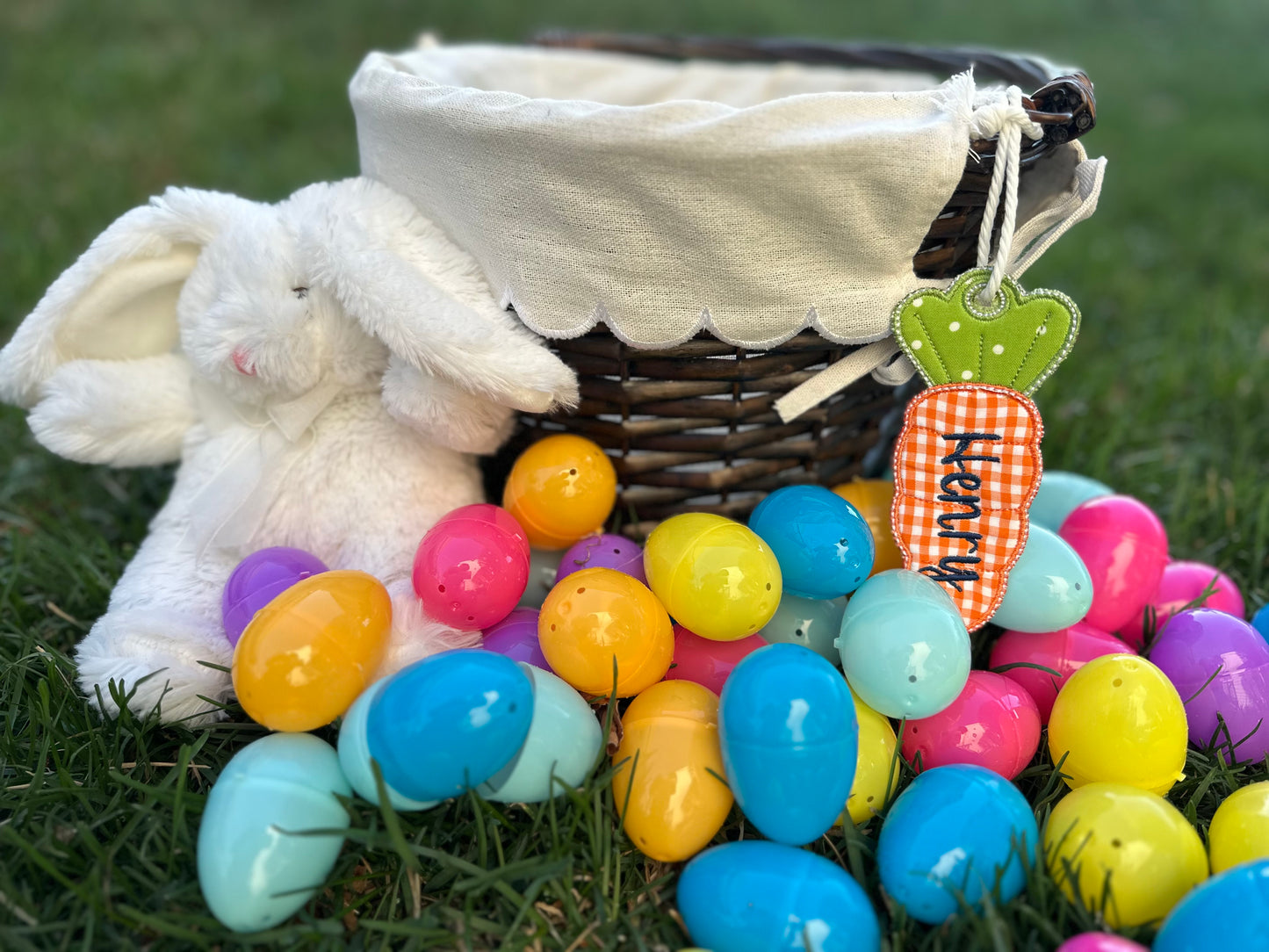 Personalized Easter basket with embroidered carrot name tag labeled 'Henry,' surrounded by colorful plastic eggs and a plush white bunny, displayed on green grass.