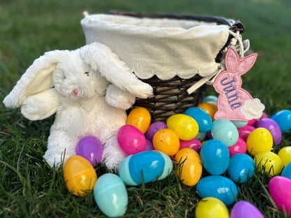 Personalized Easter basket with embroidered pink gingham bunny tag labeled 'June,' surrounded by colorful plastic eggs and a plush white bunny, displayed on green grass.