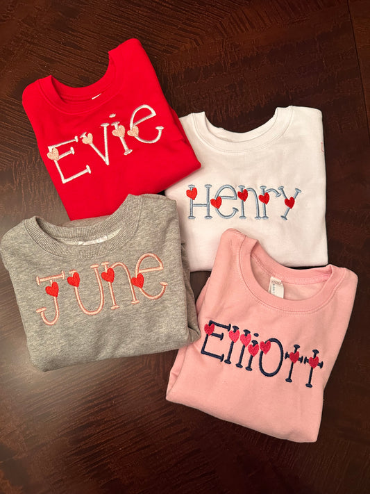 Personalized Valentine’s Day embroidered sweatshirts for kids featuring custom names with heart-themed designs, showing Evie in white lettering with light pink hearts on a red sweatshirt.  Henry in light blue with red hearts on a white sweatshirt.  June in light pink with red hearts on a grey sweatshirt.  Elliott in navy with dark pink hearts on a light pink sweatshirt.